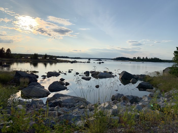 Solnedgång vid en lugn sjö med stenar i förgrunden och små öar i bakgrunden.