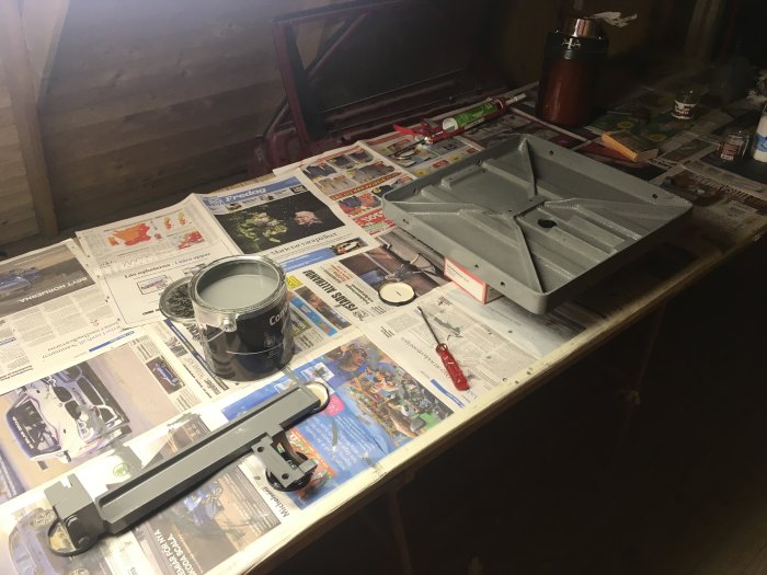 Ett arbetsbord täckt med tidningar och en gråfärgad maskindel, en burk med färg, pensel och andra verktyg.