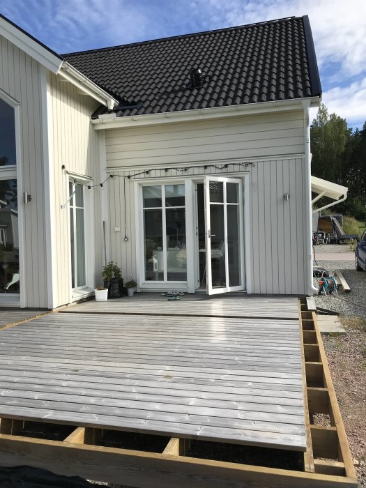 Träveranda under konstruktion vid ett vitt hus med svart tak och burspråk, oförankrad stolpe vid hörnet.