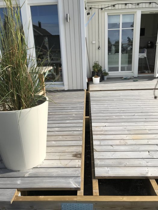 Delvis konstruerad träaltan med synliga träreglar och mellanrum, intill en husvägg med fönsterdörr och växt i kruka.