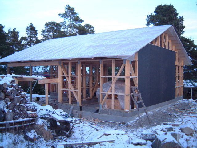 Delvis byggt trästomme till ett hus med snedsträvor och tak presenningsövertäckt i vintermiljö.