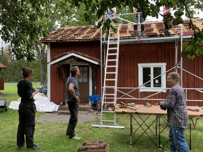 Tre personer arbetar med att lägga tegelpannor på ett rött hus medan en fjärde betraktar arbetet från marken.