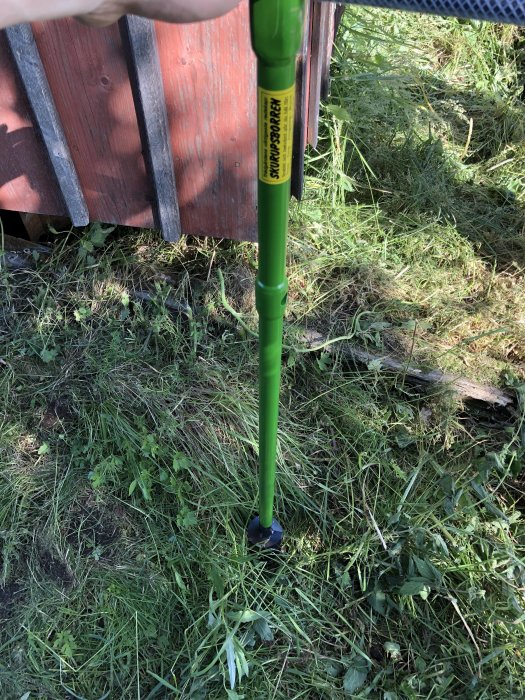 Hand håller en grön jordborr redo att borra i marken för ett provhål nära en rödfärgad stuga.