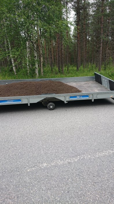 Släpvagn med tappat hjul lastad med matjord, står på en grusväg med skog i bakgrunden.