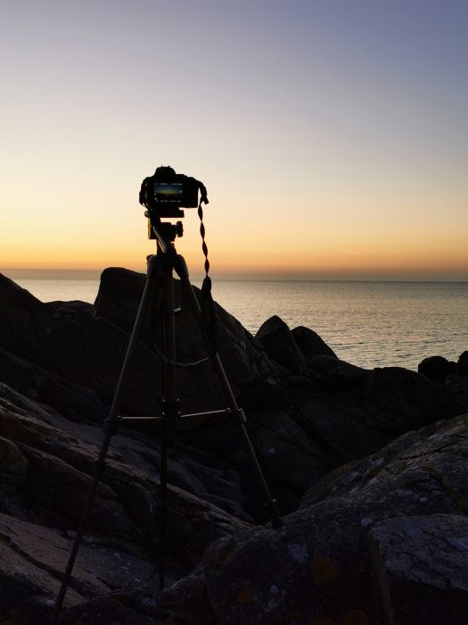 Kamera på stativ mot en solnedgång vid havet, silhuett mot en färgrik himmel.