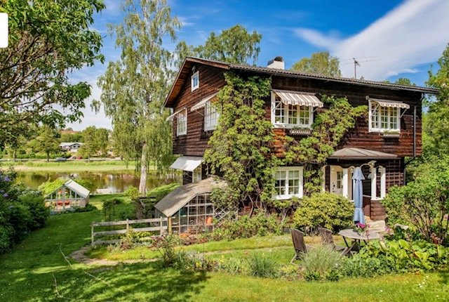 Ett tvåvånings 30-talshus med mörk träfasad, gröna fönsterluckor och en frodig trädgård.