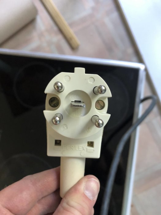 Hand håller en vit Perilex-plugg med text och specifikationer, framför ett eluttag, kabel ledande uppåt.