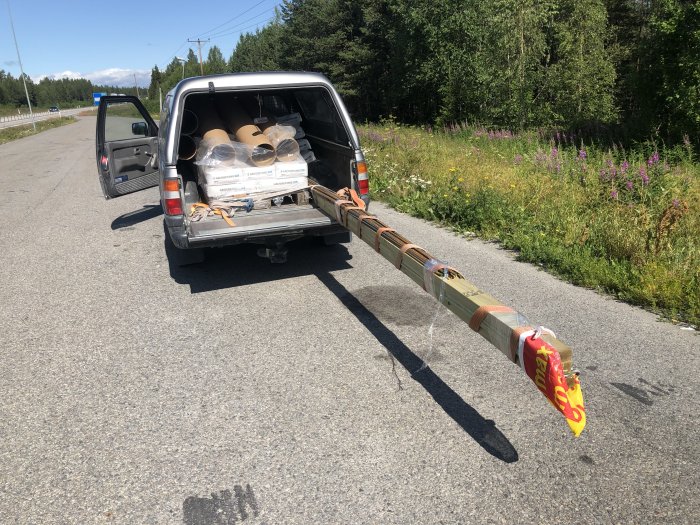 En pickup med öppen baklucka lastad med byggmaterial, längs med en landsväg.