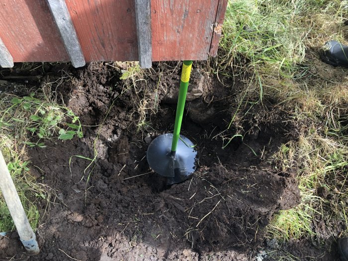 Grävningsarbete med spade för att placera sektion under hörnet på en röd byggnad, jordhög och gräs synligt.