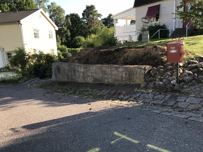 Gammal betongmur vid tomtgräns med uppgrävd del bakom och stenläggning framför, i ett villaområde.