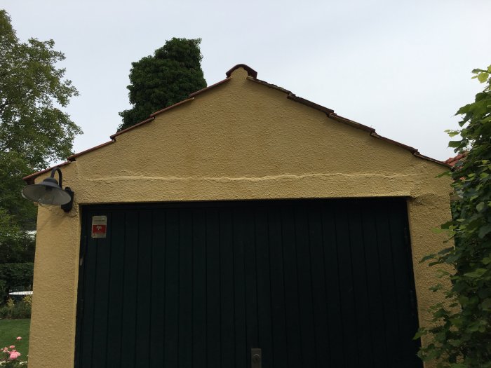 Kuva vanhasta autotallista, jossa on rapattu keltainen julkisivu ja tummanvihreä ovi. Kattorakenteessa näkyy kulumia.