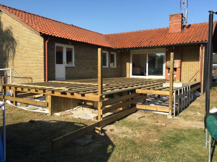 Delvis konstruerat trädäck med flera lager träbalkar framför ett gult tegelhus med röda takpannor.