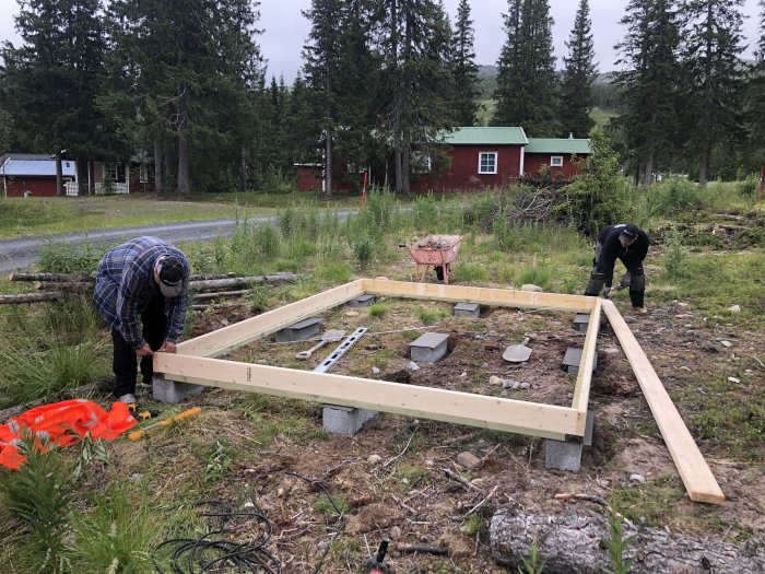 Två personer bygger en träram till ett förråd på en gräsbevuxen tomt med röda stugor i bakgrunden.