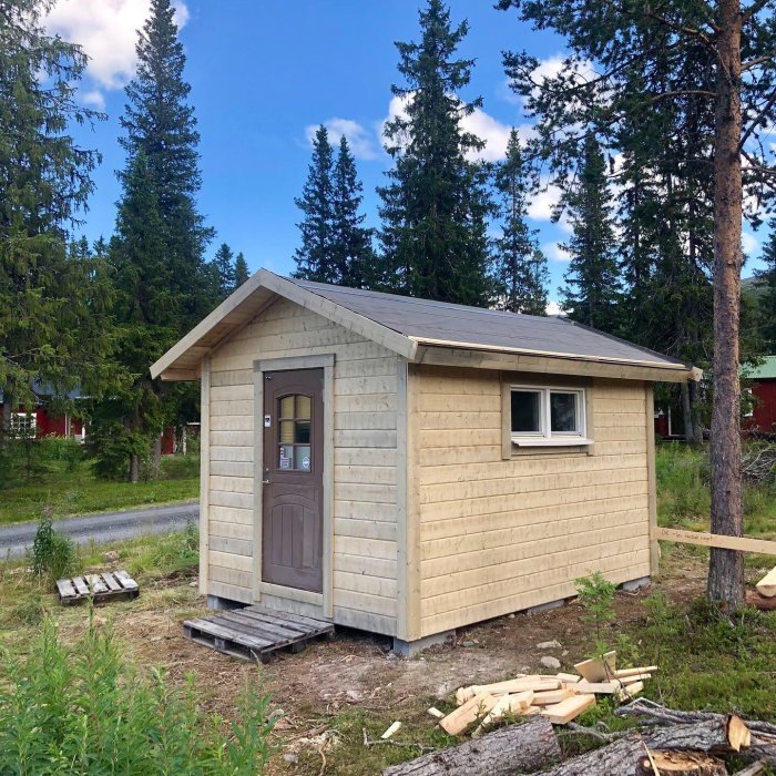 Nytt förråd på 12 kvm med ljus träpanel, återanvänt fönster och dörr, beläget i skogsmiljö.