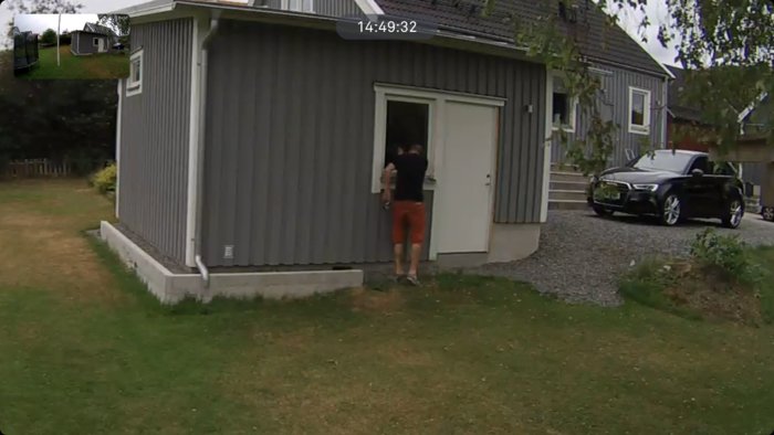 Person står vid öppen dörr till en grå uthusbyggnad bredvid en villa och en parkerad bil.