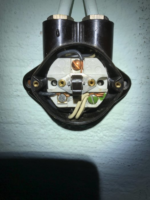 Öppet vägguttag med synliga ledningar och kopplingar, mot en målad vägg.