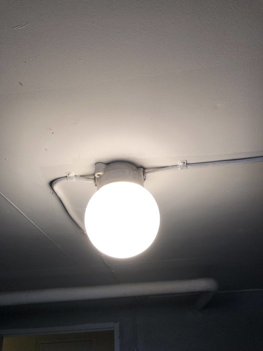 Taklampa i källaren med synlig ledning och rörinstallation mot vit takyta.