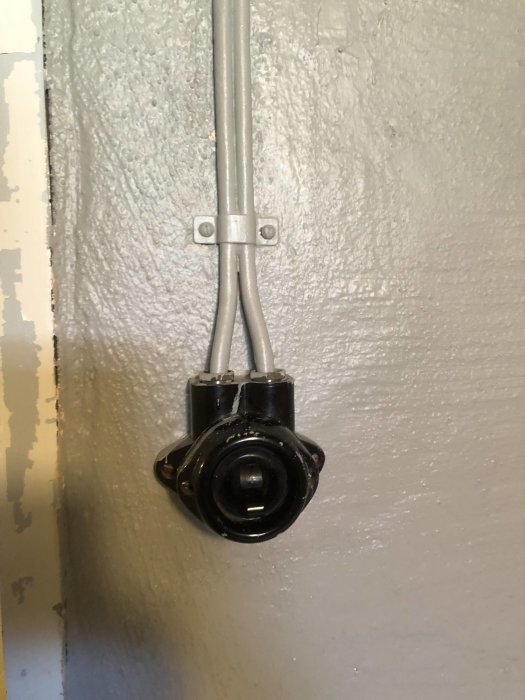 Gammal ojordad eluttag i källare med metallrör som löper neråt till ett svart uttagskropp fastsatt mot en grå vägg.
