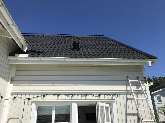 Fasad av en byggnad med vit panel och svart takpannor, stege lutad mot huset.