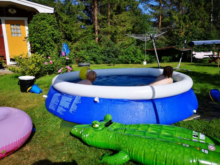 Person som sitter och vilar i en uppblåsbar pool, omgiven av uppblåsta badleksaker och grönt gräs.