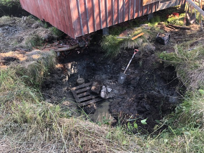 Grävd grop med vatten och lera bredvid ett hus, redskap och en pump syns.