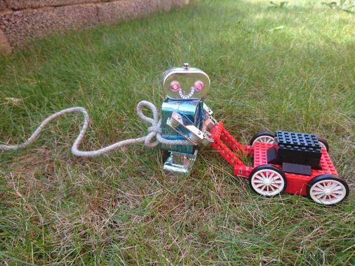 Metallisk vind-upp robot som drar en röd och svart LEGO vagn på gräset.
