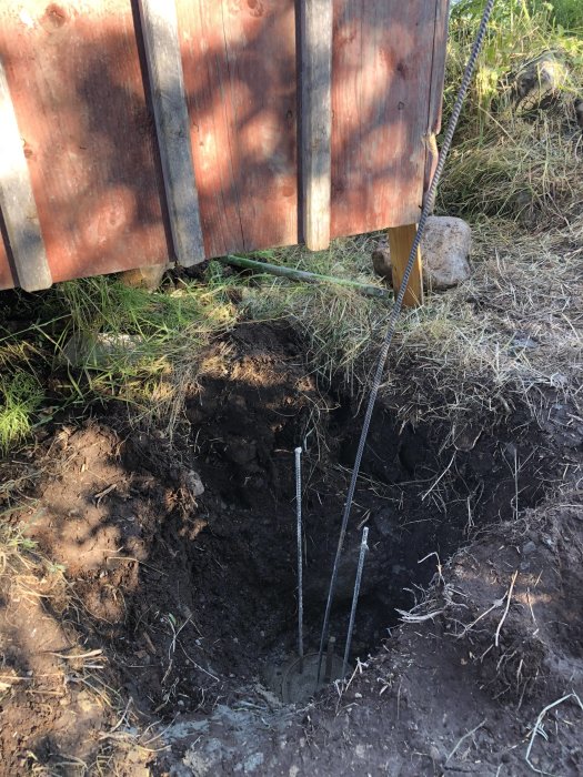 Grävt hål med armeringsjärn intill staket, packad lera och gjutrör synlig i botten.