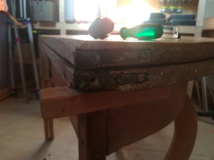 Gammalt gångjärn monterat på en träplatta, med synlig avsmalnande tjocklek och verktyg i bakgrunden.