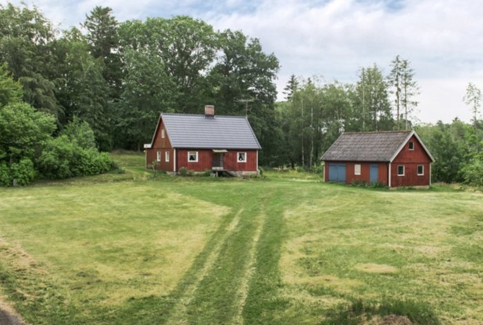 Röd villa med grått tak och tillhörande rött garage omgiven av gräsmatta och skog.