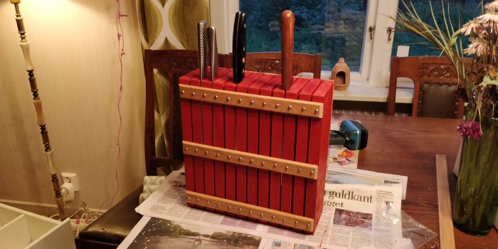 Hemmagjort knivblock av rödbetsat furuträ med eklister, fäst på ett rullbord, med knivar insatta.