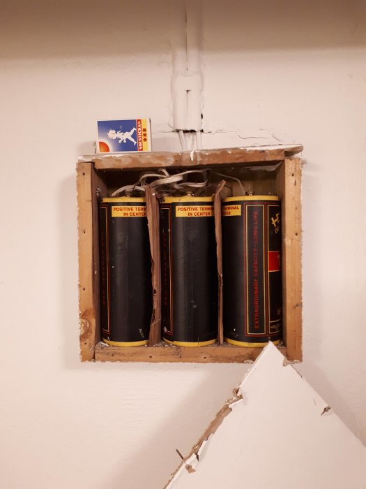 Gammal väggmonterad låda med fyra stora batterier och elkablar som går upp i taket.