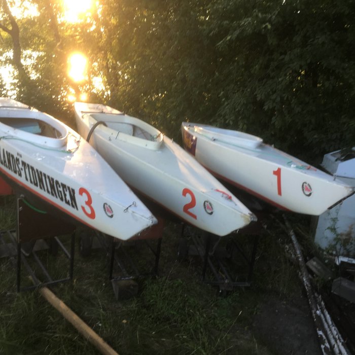 Tre vita segelbåtar numrerade 1, 2 och 3 på båtställ i grönska vid solnedgång.