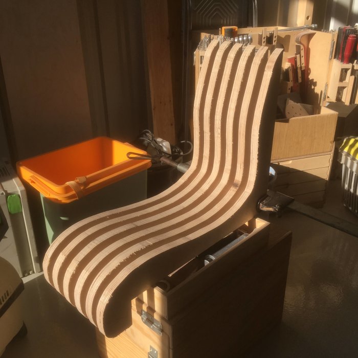 Ljussatt träarbete av ihopfogade lameller formade som en kaptensstol i en verkstadsmiljö.