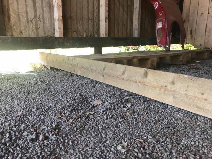 Grusgolv i carport med träbjälkar och OSB-skiva bakom, indikation på planering för gjutning av betongplatta.