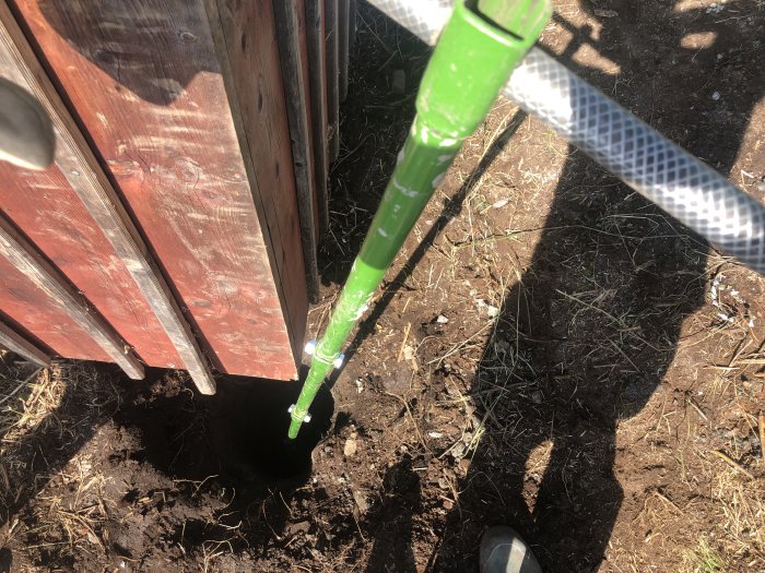 En grön förlängare för trädgårdsredskap instucken i en jordhög bredvid en trästruktur och fot.