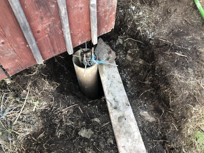 Grävt hål vid hörn av hus med sten i botten och rör delvis neddrivet i jorden.