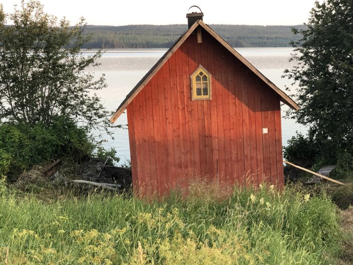 Rött trähus med lutande tak vid en sjö, synliga rörelser i husets grund indikerar små ojämnheter.
