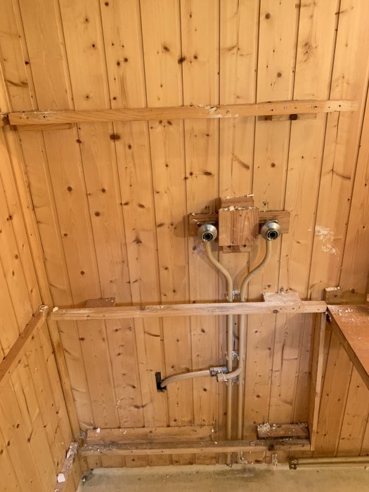 Bild av ett badrums hörn med träpanel, rördragningar och avlägsnad hemmabyggd duschkabin.