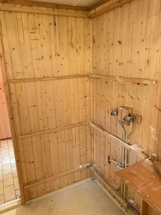 Badrumsrenovering med träpanel på väggarna och kvarvarande rör efter borttagen hemmabyggd duschkabin.