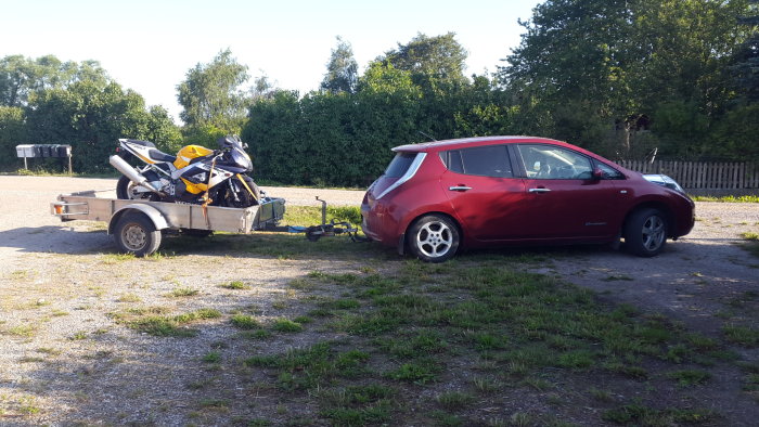 En vinröd Nissan Leaf 2012 kopplad till ett släp med en lastad motorcykel, parkerad utomhus.