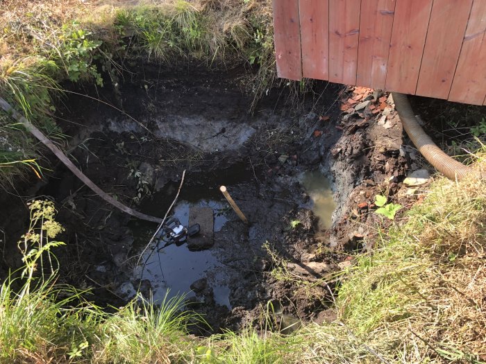 En grävd grop med vatten i botten nära en byggnad, med borrutrustning synlig, markerar avslutat borrande.