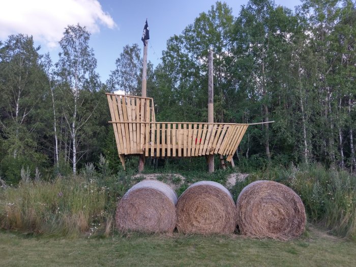 Träkonstruktion av ett skepp med trimmat räcke och synlig belysning monterad mellan två master, står på halmrullar.