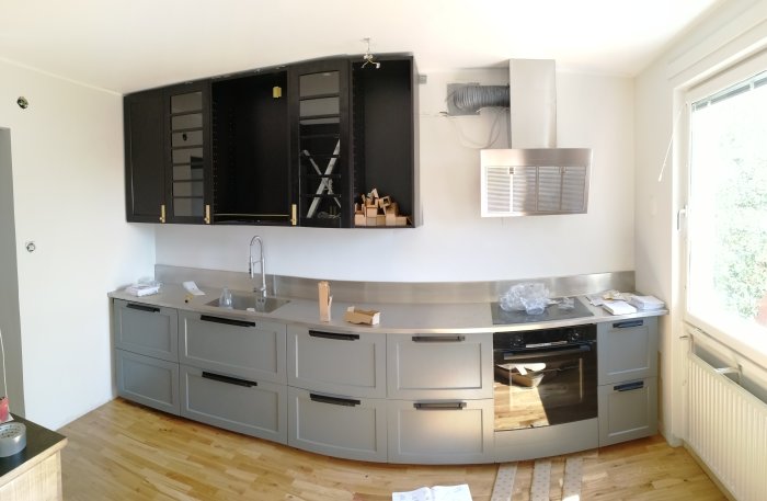 Kök i renovering med grå skåpsluckor, öppet skåp utan dörr, rostfri bänkskiva och trägolv.