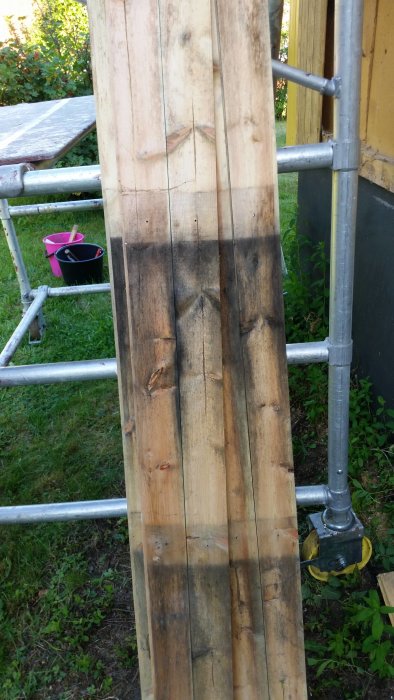 Träpanel med tecken på svartmögel och fuktskador, med byggställning och trädgård i bakgrunden.