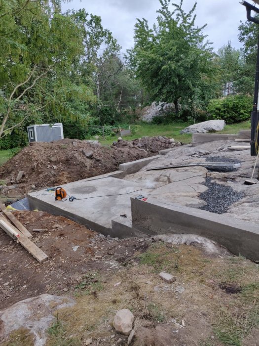 Nyggjuten betongsockel och platta för bergvärmepump under bygget av ett hus, med verktyg och trädgård i bakgrunden.