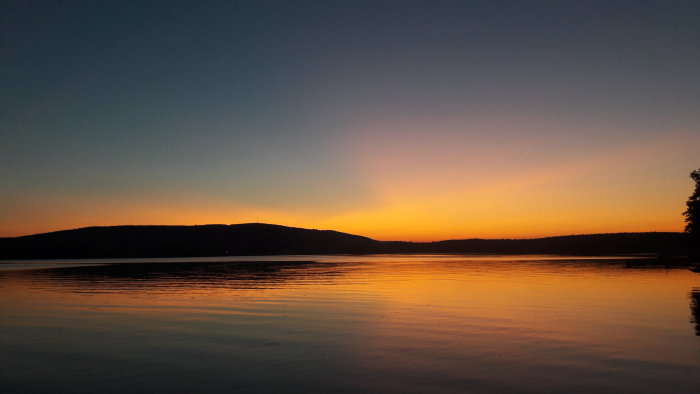 Solnedgång med orange och blå himmel speglande i en stilla sjö med siluett av kullar i bakgrunden.