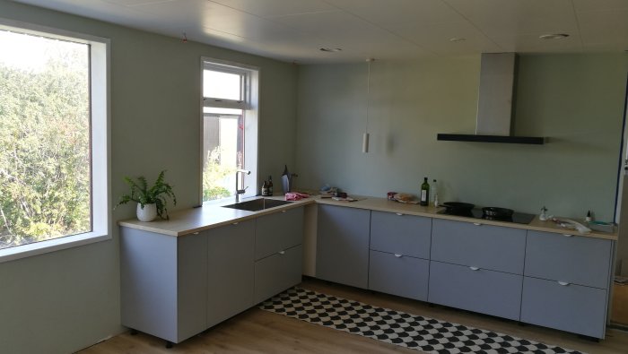 Kök under renovering med gråa skåp, vit bänkskiva och svartvit rutig matta.