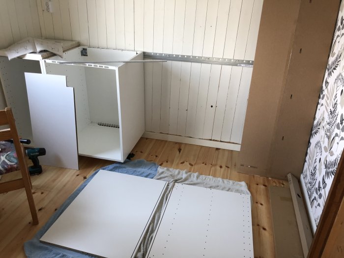 Omonterade vita köksskåpsdelar och en verktyg spridda på ett trägolv under renovering.