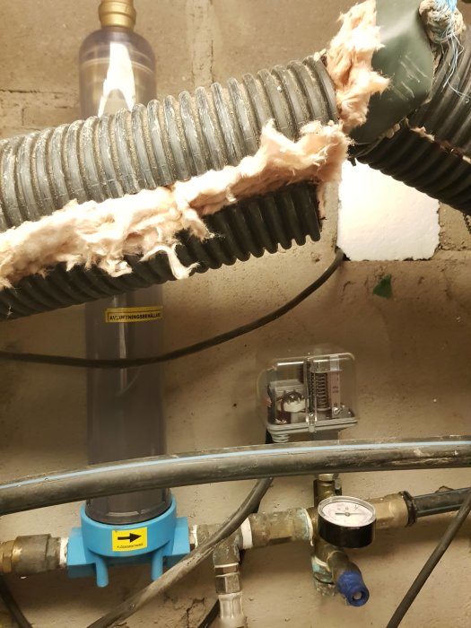 Tryckmätare på en vattenledning i en källare med fast visare som visar 0.2 bar, omgiven av rör och isolering.