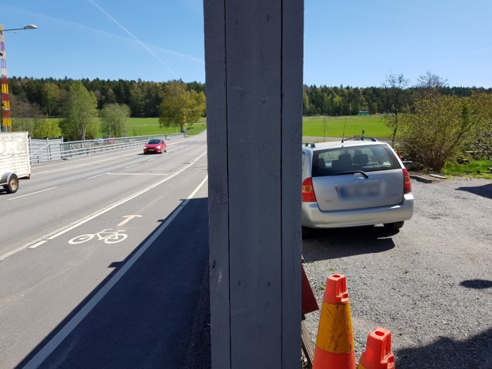 Bullerplank av trä sett nära en bilväg med en parkerad bil och trafikkoner i förgrunden.